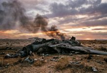 Military jet aircraft wreckage with smoke in desert landscape