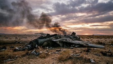 Military jet aircraft wreckage with smoke in desert landscape