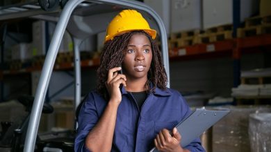 focused-black-warehouse-worker-yellow-hardhat-standing-near-forklift-talking-cell-shelves-with-goods-background-medium-shot-labor-communication-concept-1